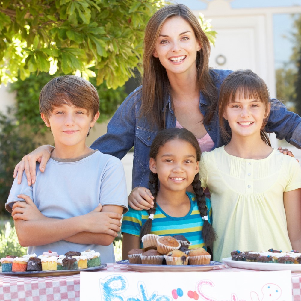 Group of children holding bake sale with mother smiling to camera.